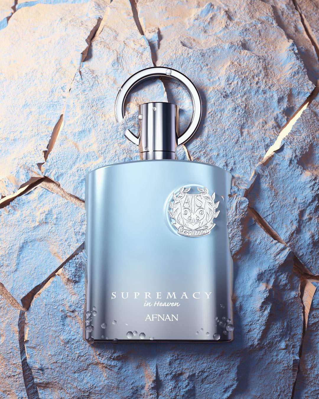 A bottle of Afnan's "Supremacy in Heaven EDP Perfume for Men 3.4 oz/100 ml" rests on a cracked, light blue stone surface, evoking a tranquil atmosphere. This light blue bottle with silver accents features a round decorative emblem and a metallic loop-like cap, while the brand name "AFNAN" is elegantly displayed at the bottom.