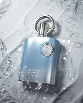 A sleek blue bottle of Supremacy in Heaven EDP Perfume for Men by Afnan, containing 3.4 oz (100 ml) of fragrance, is surrounded by splashes of water, evoking a tranquil atmosphere. The bottle features a metallic cap and an intricate emblem on its front, set against a textured light grey background.