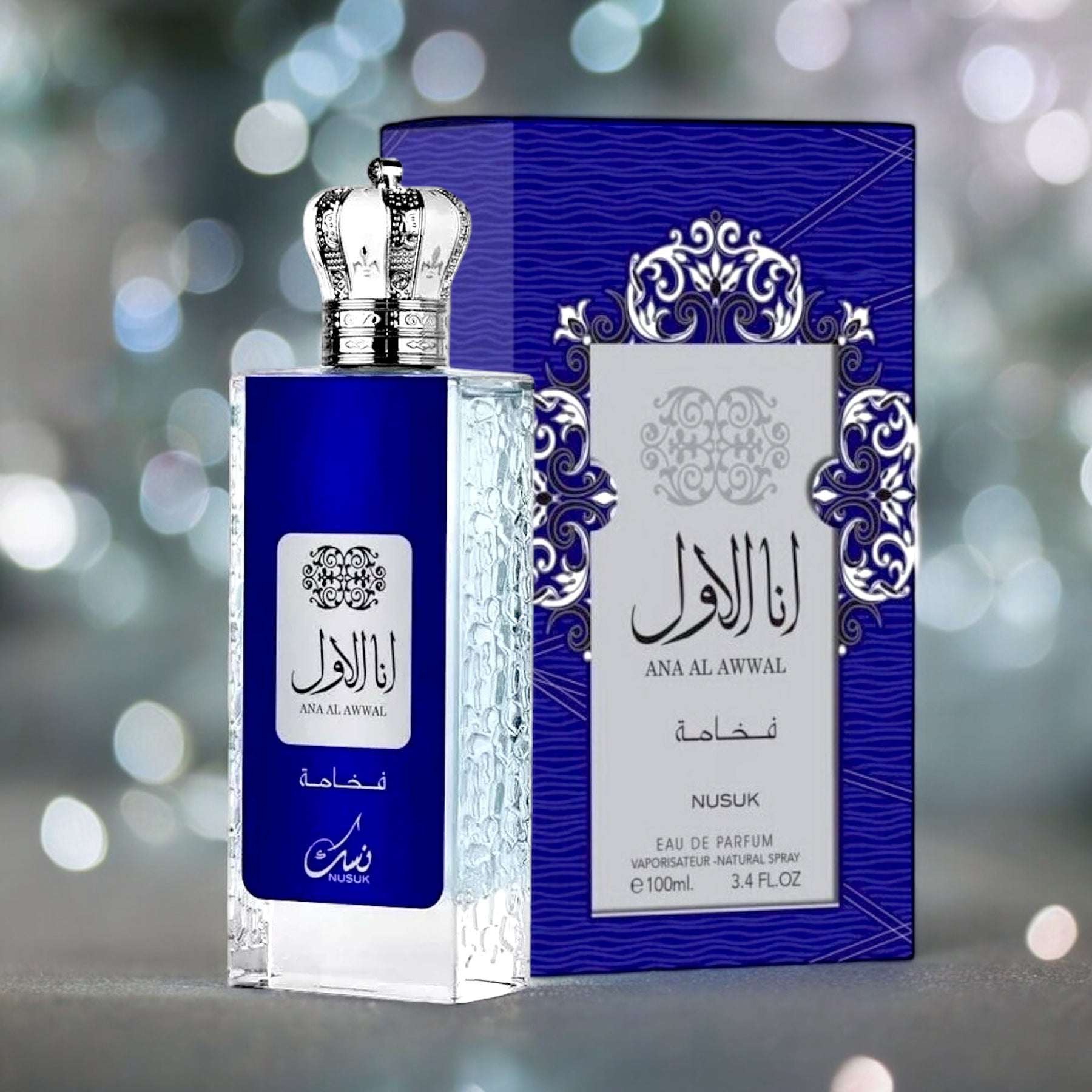 A blue 3.4 oz/100 ml perfume bottle named "Ana Al Awwal Blue EDP Perfume for Men" by Nusuk, featuring a silver cap, is placed beside a coordinating blue and silver ornate box. Both the bottle and the box are adorned with Arabic script. The background is softly blurred with white and green bokeh lights, adding to the ambiance. The text on the bottle reads "Ana Al Awwal - Nusuk," highlighting its versatility and long-lasting香 notes.