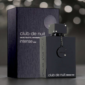 An Armaf "Club de Nuit Intense Man EDT Perfume for Men" bottle, boasting a sleek black design and holding 3.6 oz (105 ml) of woody spicy fragrance that embodies modern masculinity, stands proudly in front of its matching black box packaging. Both the bottle and the box feature white text highlighting the product's name. The background is artistically blurred with light spots.