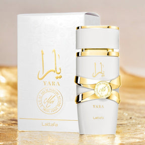 A white and gold perfume bottle labeled "Yara Moi EDP Perfume for Women 3.4 oz/100 ml" by Lattafa is displayed in front of its matching packaging, showcasing an elegant design with Arabic script and branding. Crafted for sophisticated women, the softly blurred background features golden hues.